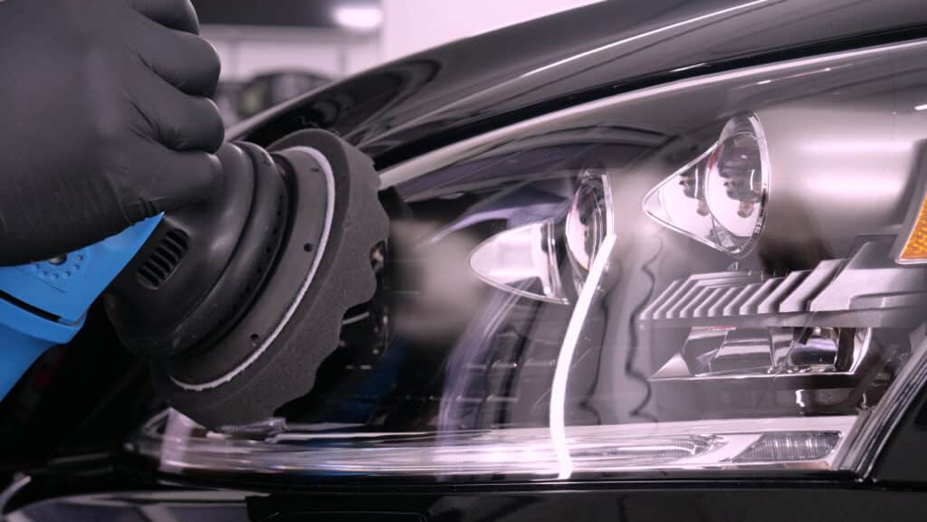 Main gantée polissant le phare d'une voiture noire avec une machine de detailing bleue, restaurant la clarté et la brillance.