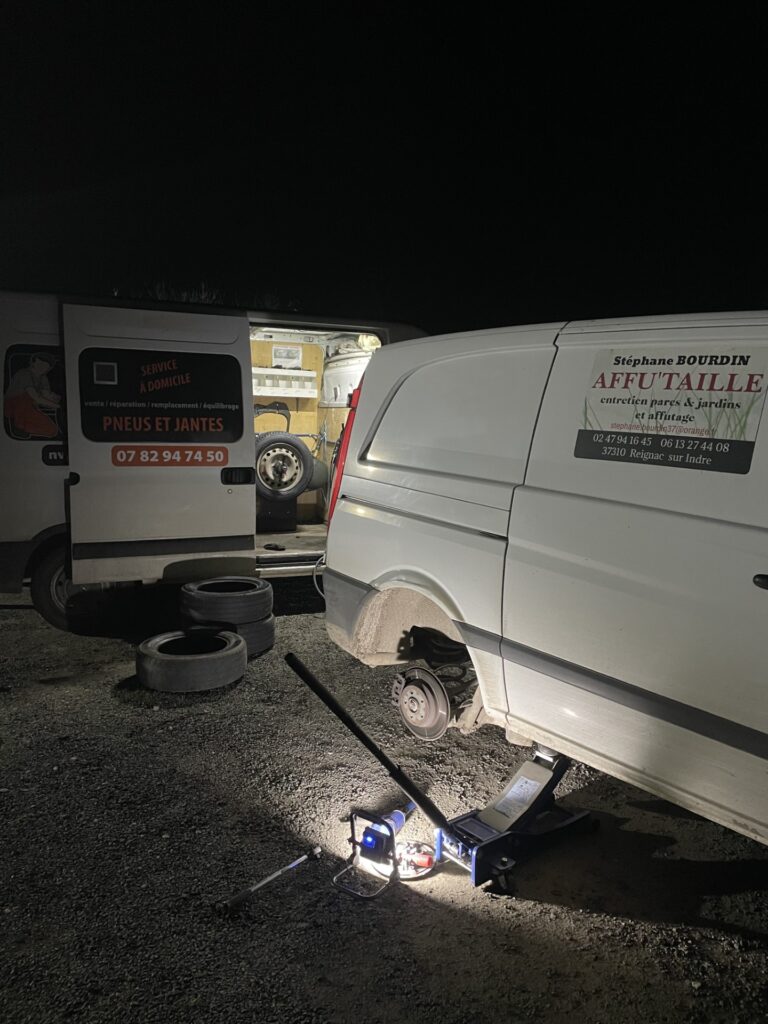 Service mobile de pneus la nuit. Van 'PNEUS ET JANTES' répare roue sur cric, pneus au sol. Van 'AFFU'TAILLE'.