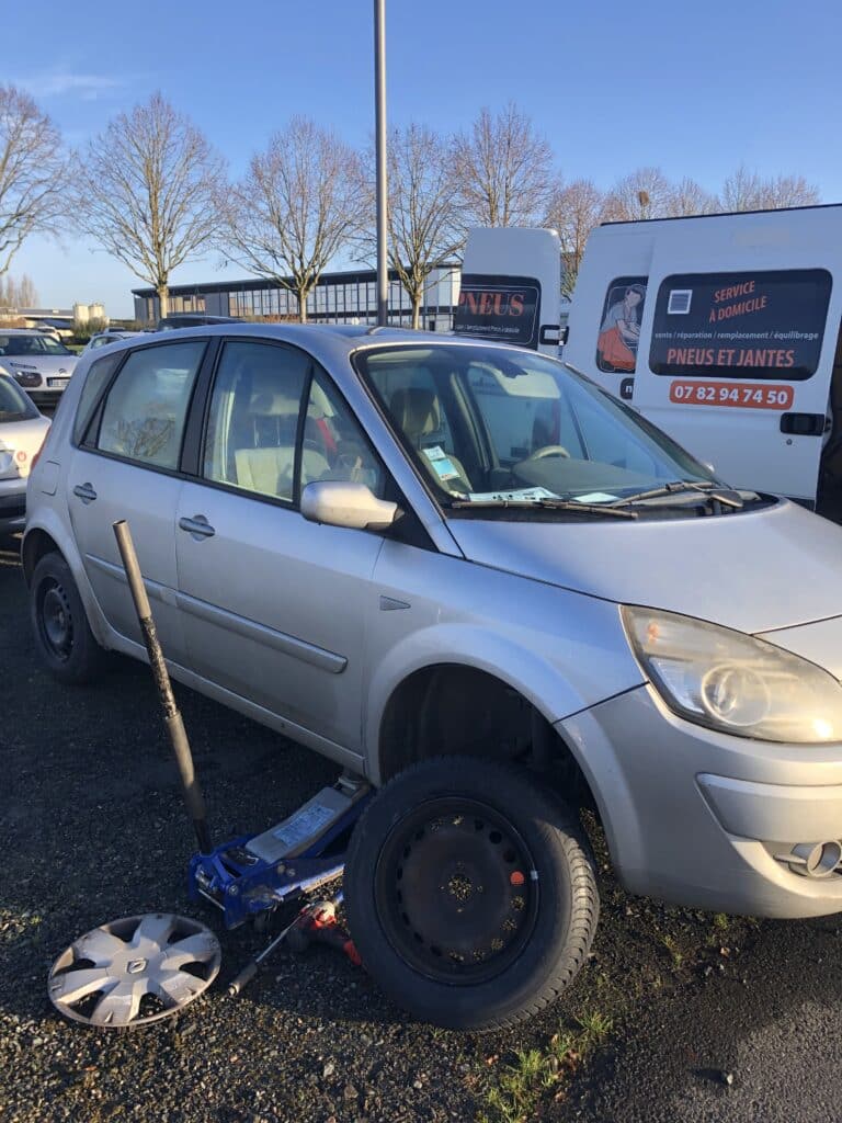 Voiture argentée sur cric, roue avant retirée, près d'un van "PNEUS ET JANTES" (07 82 94 74 50) offrant service de pneus à domicile.