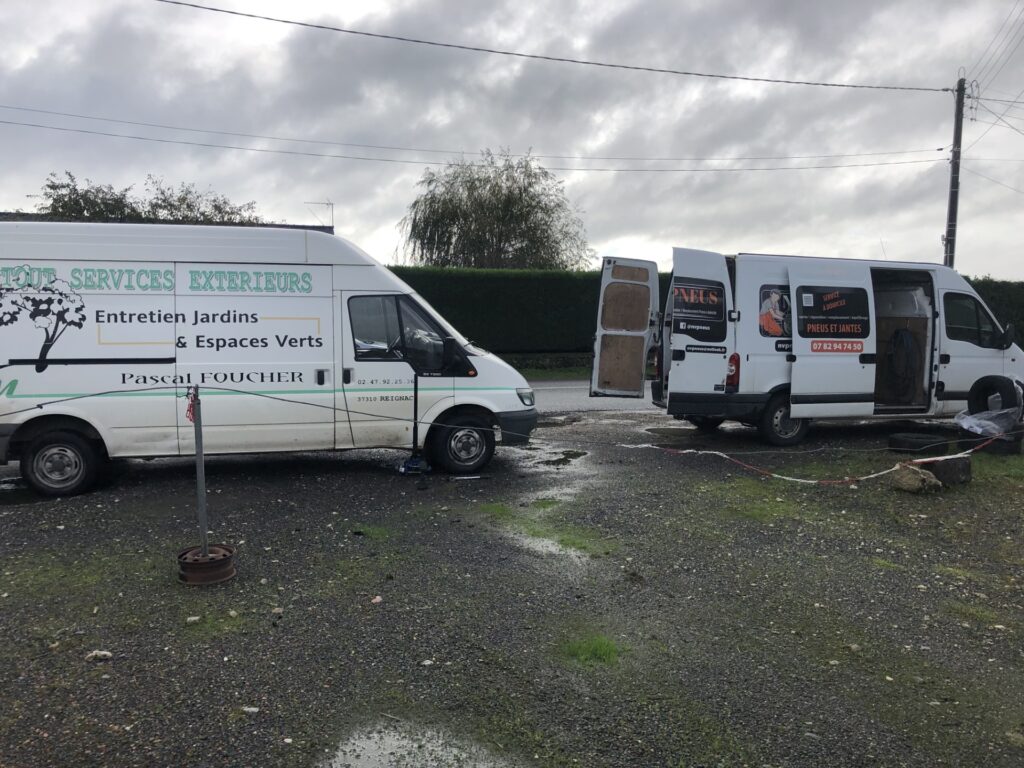 Deux fourgons de service blancs: Pascal Foucher (jardinage) et MVPneus (pneu mobile). Logos et numéros visibles. Terrain gris par temps nuageux.