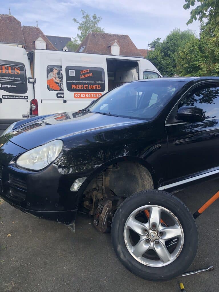 Porsche noire sans roue avant, son pneu retiré au sol. Deux vans NVPneus mobiles derrière, offrant un service de réparation à domicile.