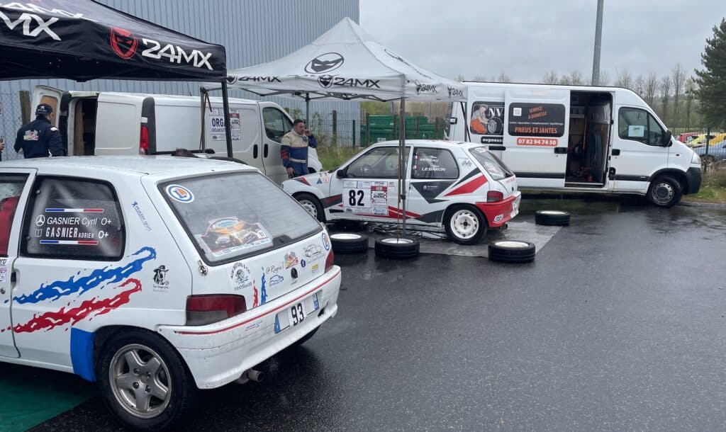 Deux Peugeot 106 de rallye blanches (numéros 82, 93) sous des tentes 24MX. Des vans de support NVPNEUS sont visibles. Un homme fume sur l'asphalte mouillé.