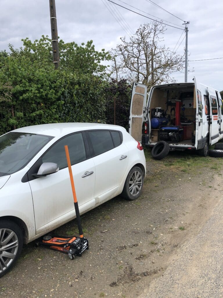 Van de dépannage pneu mobile avec équipement. Une voiture blanche est levée par un cric. Texte: PNEUS ET JANTES, GOOD YEAR.