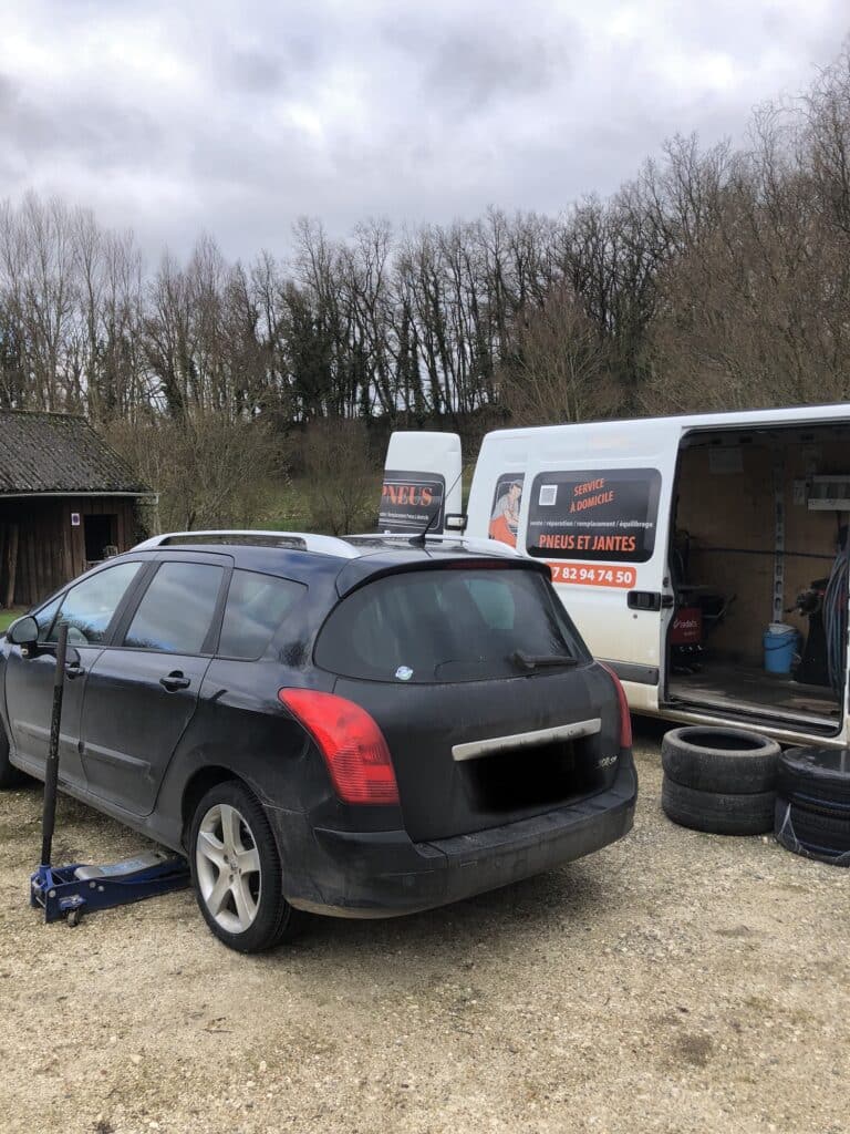 Véhicule noir Peugeot 308 SW levé sur cric, devant un fourgon blanc "SERVICE À DOMICILE PNEUS ET JANTES 07 82 94 74 50" pour réparation.